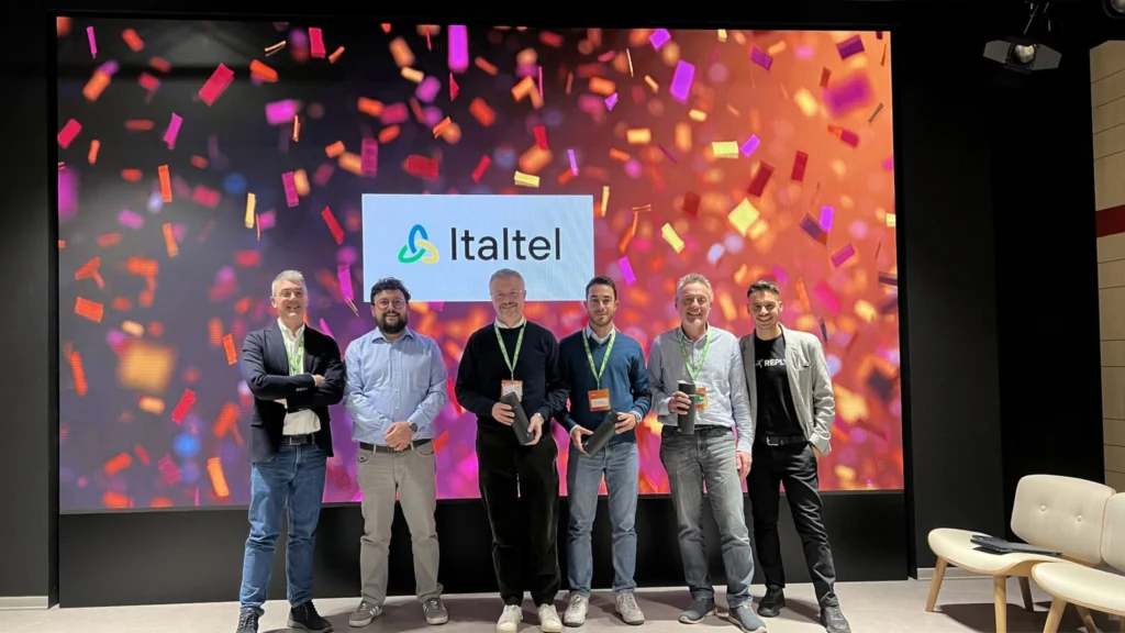 A group of six men stands smiling in front of a vibrant screen displaying colorful confetti and the Italtel logo, conveying a celebratory mood.