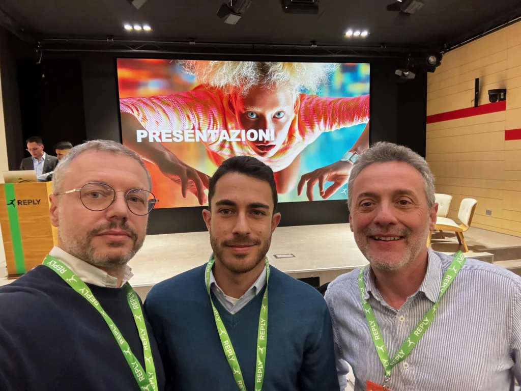 Three men with lanyards smile for a selfie in front of a large screen displaying "PRESENTAZIONI" and a dynamic image of a person. The setting is professional and vibrant.