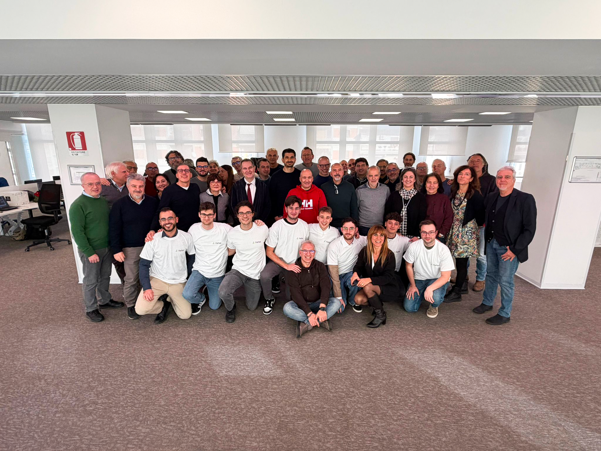 A large group of people, diverse in age and gender, smile in an office setting. They're casually dressed, conveying a joyful and collaborative atmosphere.
