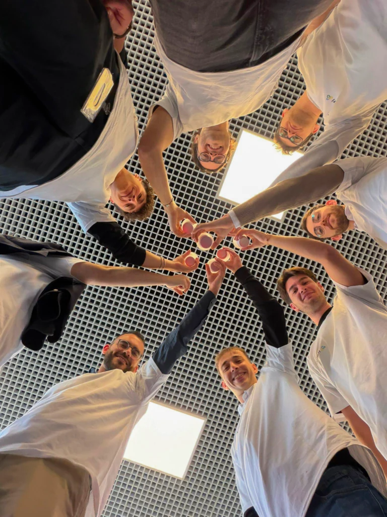 Seven people in casual white shirts form a circle and raise red cups in a celebratory pose, viewed from below against a patterned ceiling. The mood is joyful.