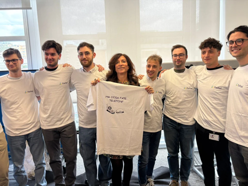 A group of eight people stand in a well-lit room, all smiling. Seven men and one woman wear matching white T-shirts. The woman holds another T-shirt with a cartoon of a phone and Italian text. The mood is cheerful and collaborative.