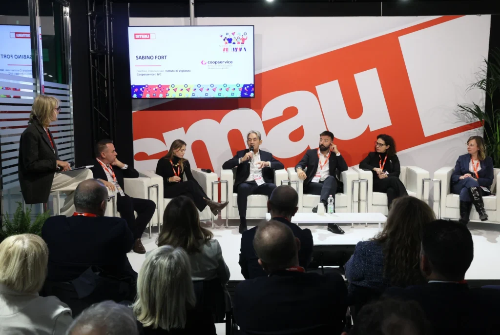 Panel discussion at a conference with six speakers on stage, seated in white chairs. A large "Smau" logo is displayed behind them. Audience visible in foreground.