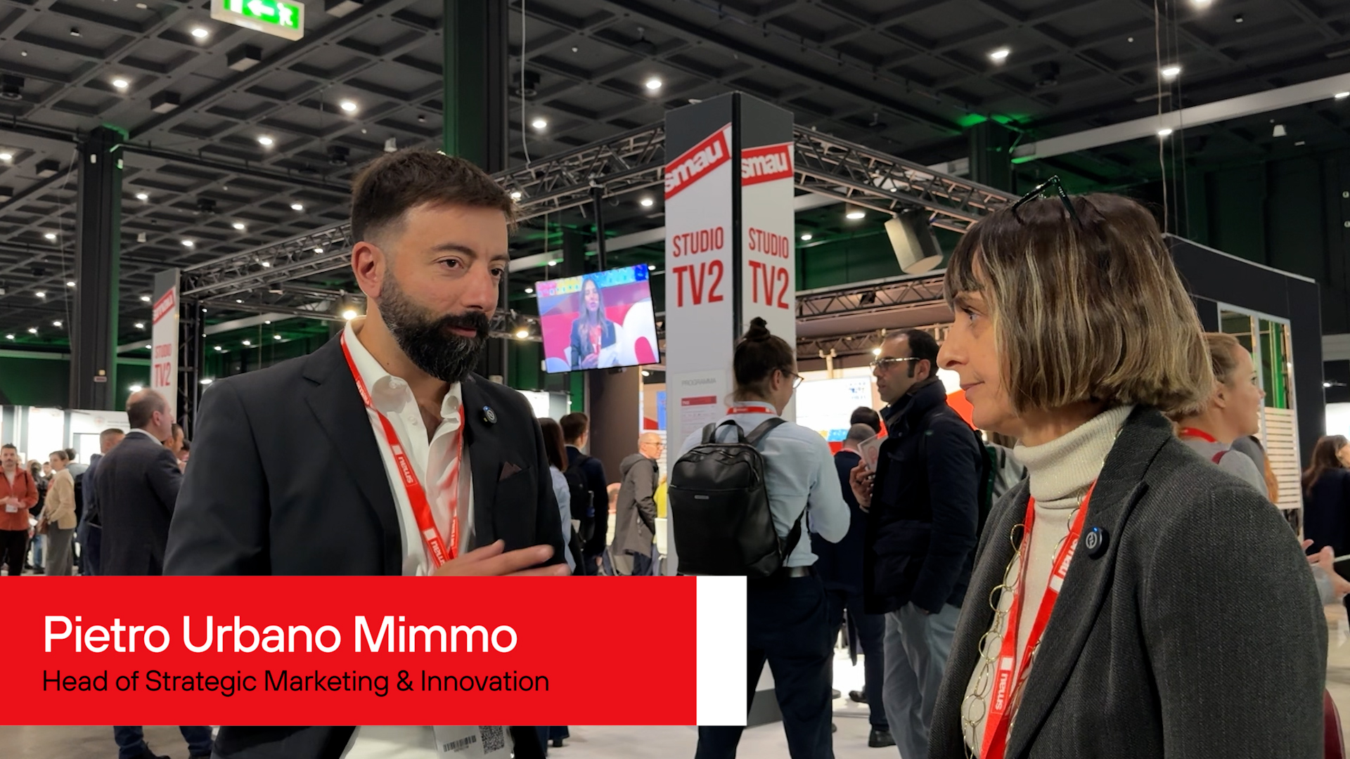 A man and a woman are engaged in conversation at a bustling conference. Both wear badges, suggesting a professional setting. The mood is focused and lively.