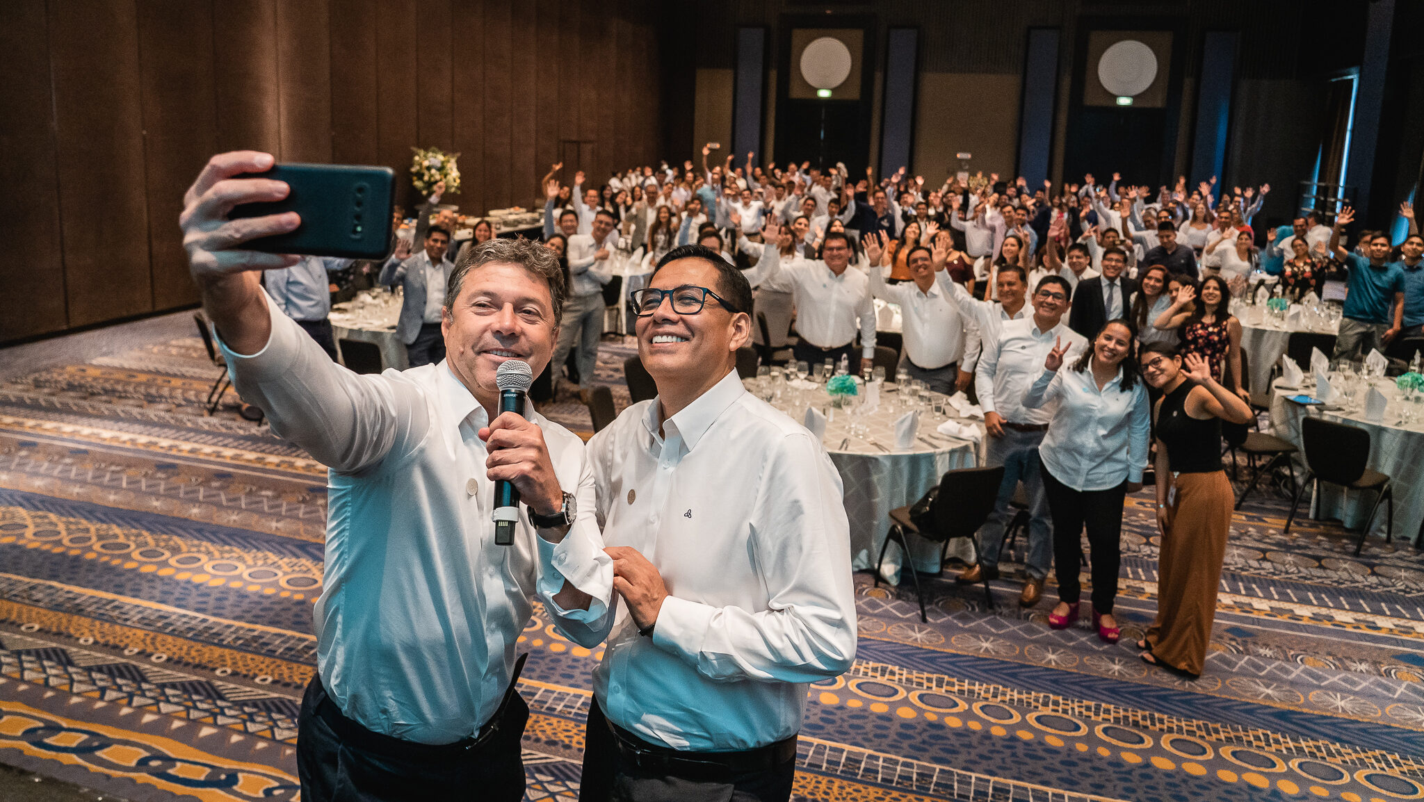 Two men of italtel peru in white shirts take a selfie with a smartphone at a large conference. Italtel team behind them smiles and waves in a spacious, well-lit hall.