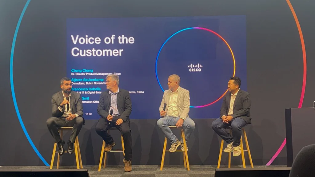 Four men sit on stools discussing on stage against a screen reading "Voice of the Customer" with the Cisco logo. The setting is professional and engaging.