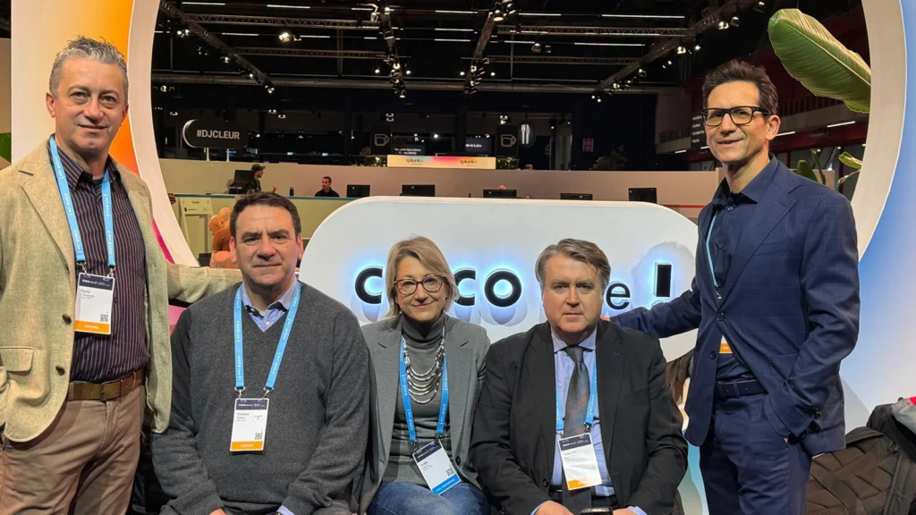 Five people are posing in front of a display at a conference. They are smiling, dressed in business attire, and wearing conference badges, suggesting a professional atmosphere.