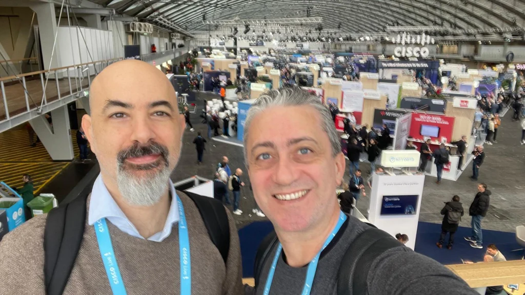 Two people smiling in the foreground of a bustling conference hall filled with booths and attendees. The ambiance is lively and engaging.