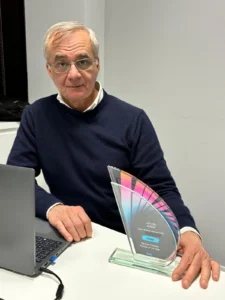 A man with glasses and a navy sweater sits at a table, hand resting on a colorful, triangular award. A laptop lies open nearby.