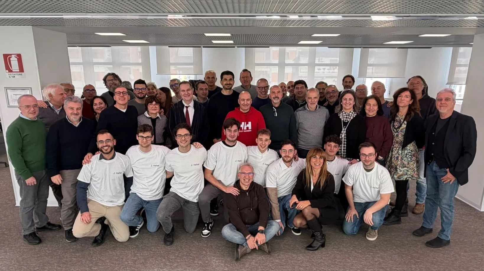 A large group of people, diverse in age and gender, smile in an office setting. They're casually dressed, conveying a joyful and collaborative atmosphere.