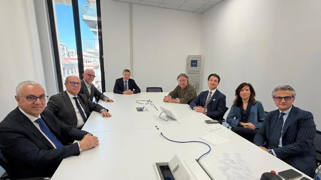 People in formal attire sit around a white conference table in a meeting room with large windows. The atmosphere is professional and collaborative.