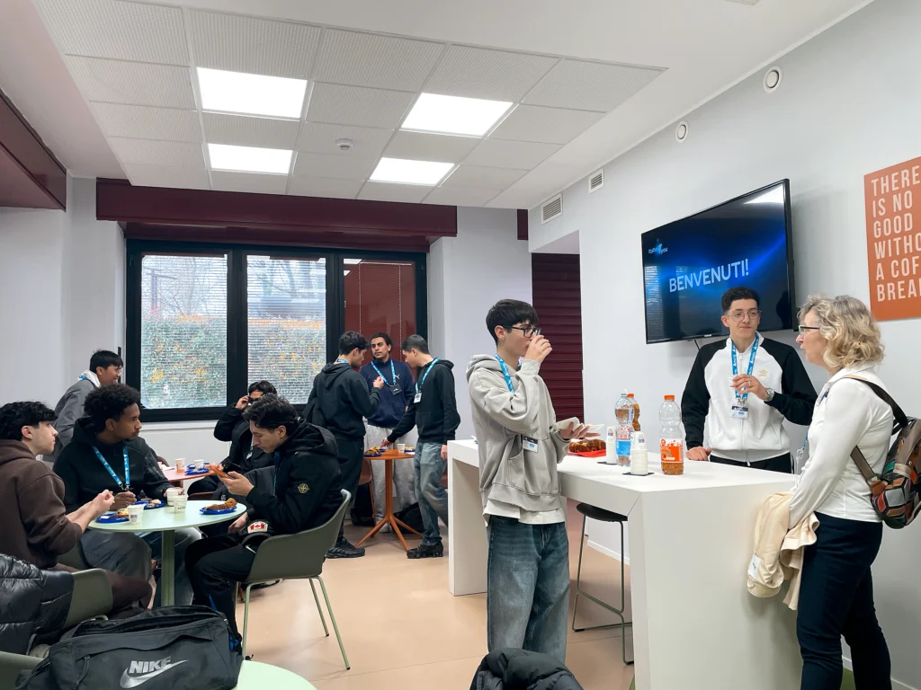 A group of people in the Italtel Coffee Break engage in casual conversation and snacks. A screen displays "Benvenuti!" in Italian. The atmosphere is friendly.