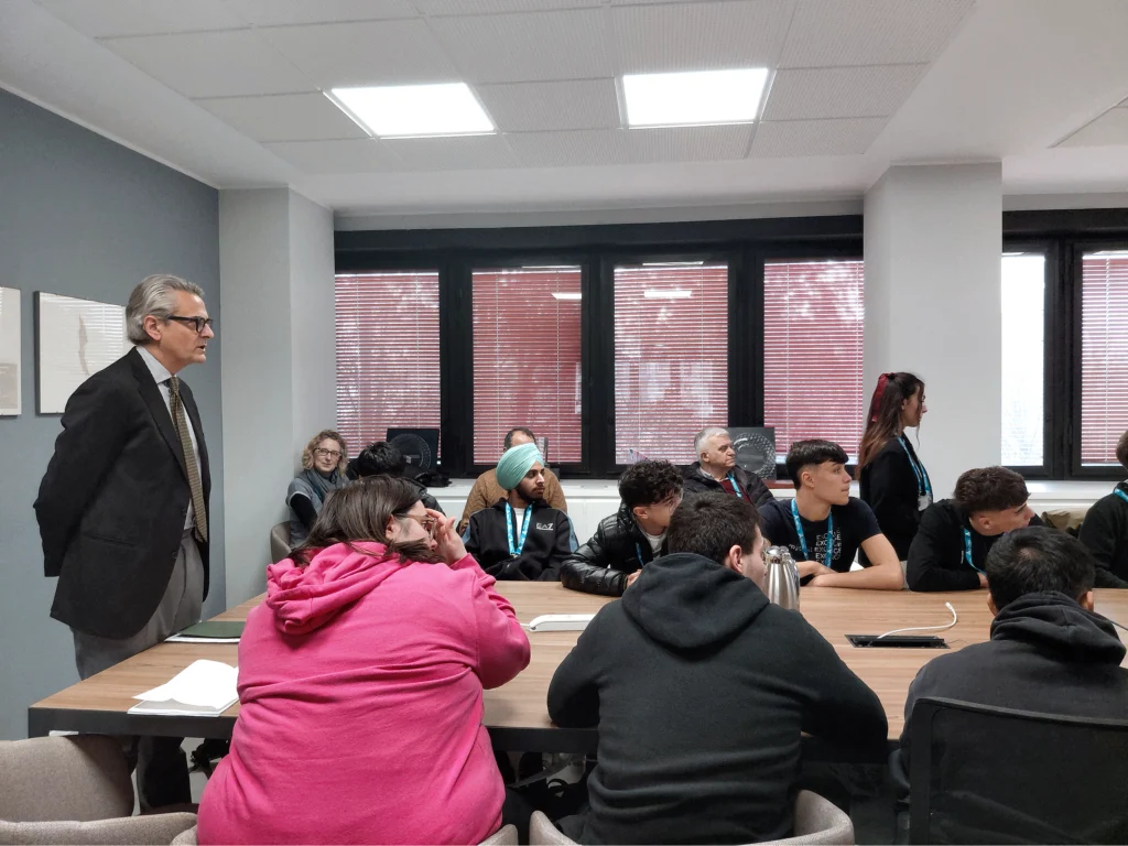A group of people sit attentively around a conference table in an office, while Michele Saracino, HR director, stands to the side, observing. The mood is focused.