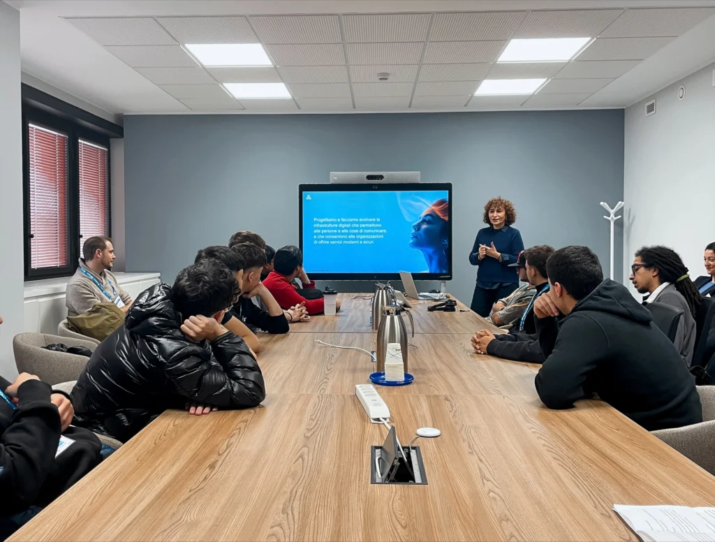 A group of people in a Italtel conference room watch a presentation on a screen. Laura Borlenghi, Head of Communication, stands near the screen, speaking, while attendees listen intently.