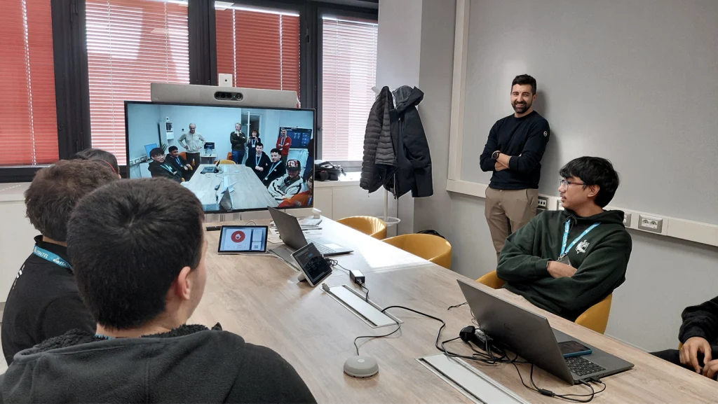 A team of five people sits around a conference table with laptops while conducting a video call on a large screen. Luca Simonini, head of competence center of Italtel, smiling by the wall.
