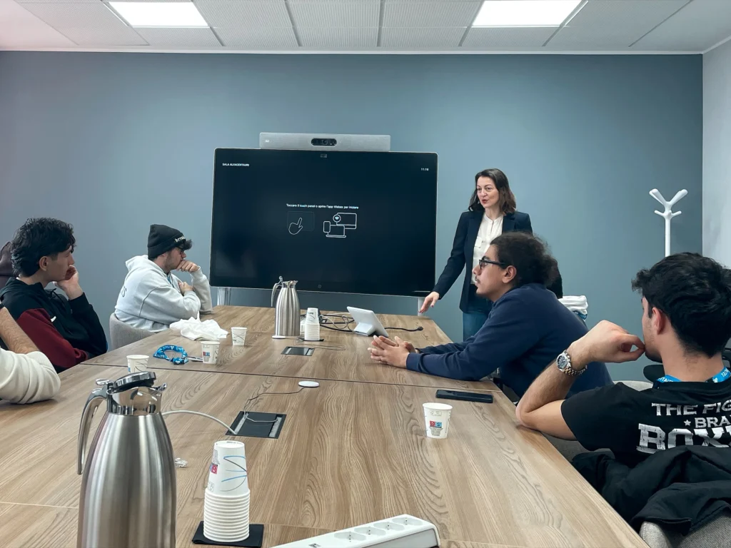 Federica Bolognesi stands by a large screen presenting to a group of attentive students in a conference room. The mood is focused and professional.