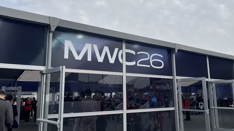 Exterior view of a tech event entrance with "MWC26" displayed prominently. A crowd is visible inside, evoking a sense of bustling activity and anticipation.
