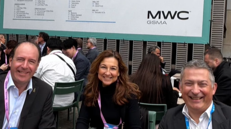 Three people smiling at an outdoor event with "MWC GSMA" signage behind them. They appear relaxed and happy, seated among others.