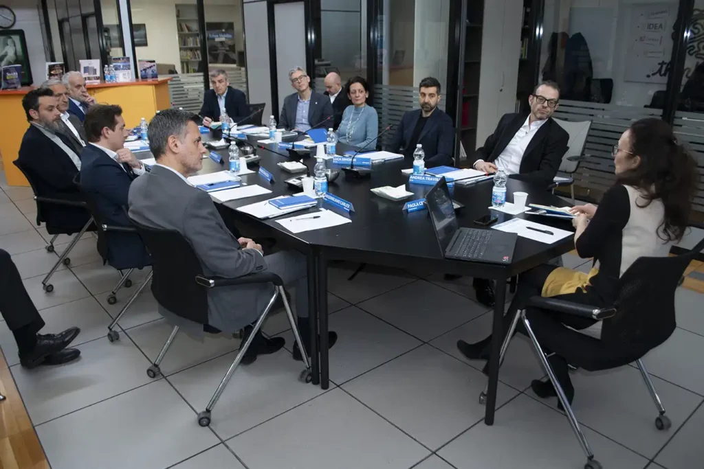 A group of professionals in formal attire seated around a large, black conference table, engaged in discussion. The room has a serious tone.