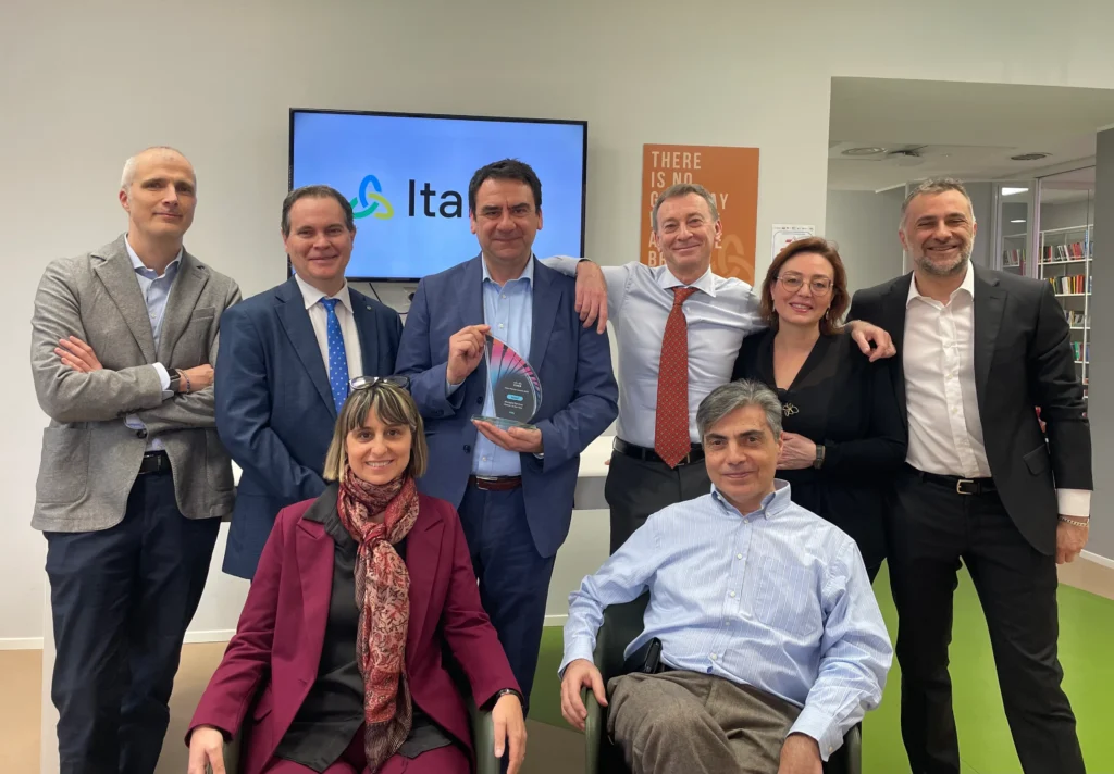 eight Italtel colleagues pose in an office, one holding an award. They smile warmly, conveying a sense of achievement and camaraderie.
