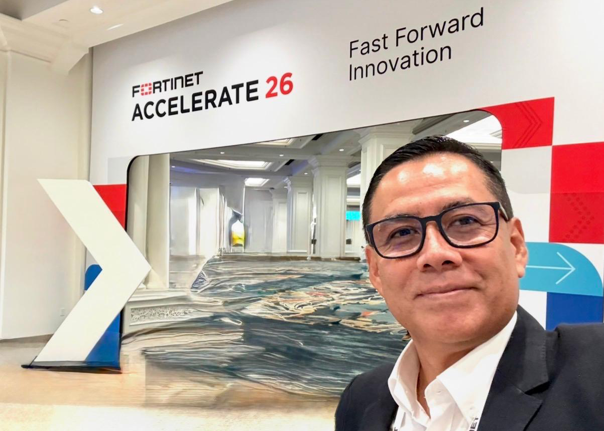 William Gonzales, Country manager of Peru & Colombia, smiles at a conference with a sign reading "Fortinet Accelerate 26, Fast Forward Innovation" in a modern, bright hallway.