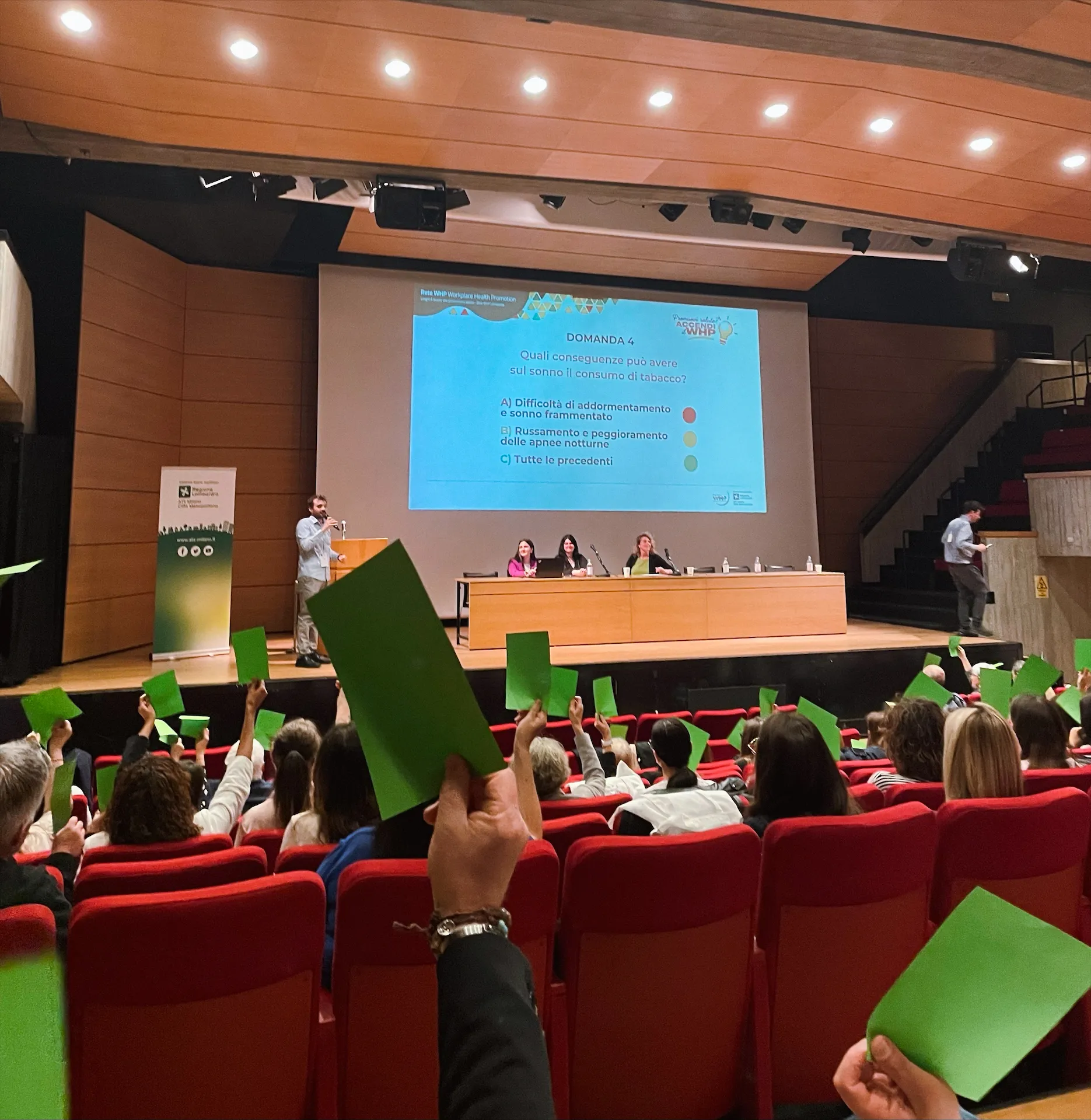 Conferenza in un auditorium: il pubblico seduto su poltrone rosse alza cartoncini verdi durante una votazione, mentre sul palco un relatore parla accanto a un grande schermo con una domanda sul sonno e il consumo di tabacco; tre relatrici sono sedute a un tavolo