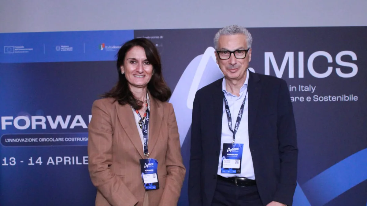 Two colleagues from Italtel (Maria Rosa Pizzo & Antonino Albanese) standing in front of a dark blue conference banner. Both wear lanyards and formal attire, smiling warmly. The mood is professional and friendly.