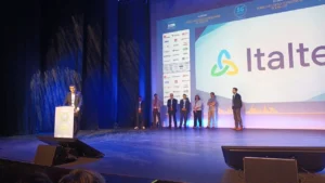 A person speaks at a podium on a stage with a large screen displaying the Italtel logo. Several people stand onstage, suggesting a formal event.