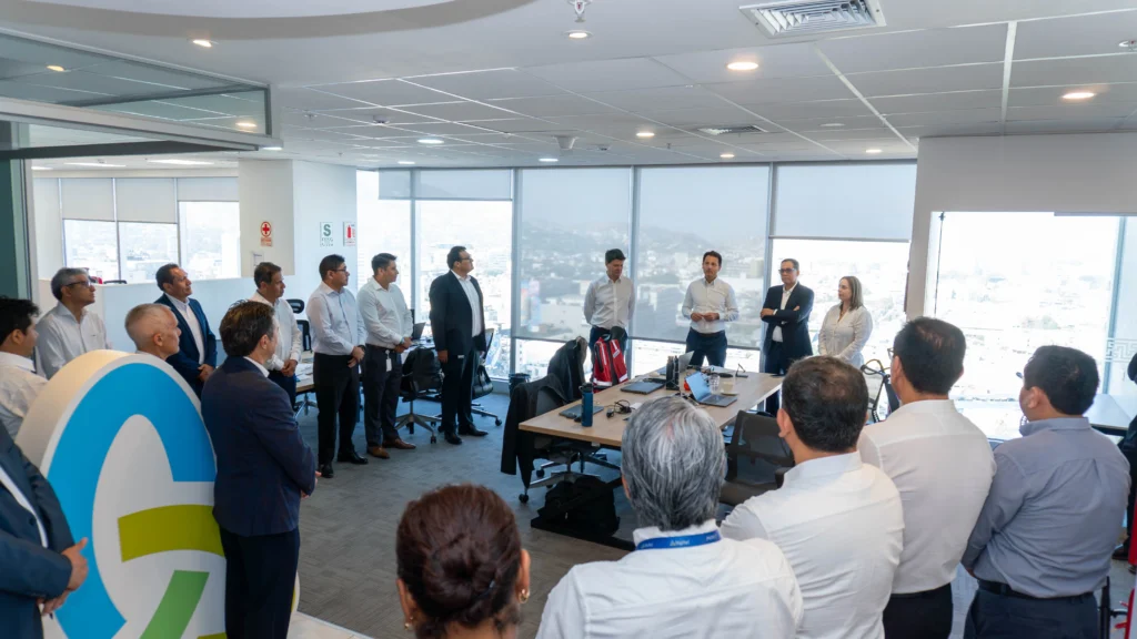 Modern corporate office meeting with a diverse group of business professionals standing in a circle during a team briefing or executive presentation. Bright open-plan office conference room with large floor-to-ceiling windows, city skyline view, laptops and office desks visible. Men and women in business attire listening to a speaker during a workplace collaboration session, corporate leadership meeting, team-building discussion, or company strategy presentation. Contemporary office interior with natural daylight, professional networking environment, and collaborative business atmosphere.