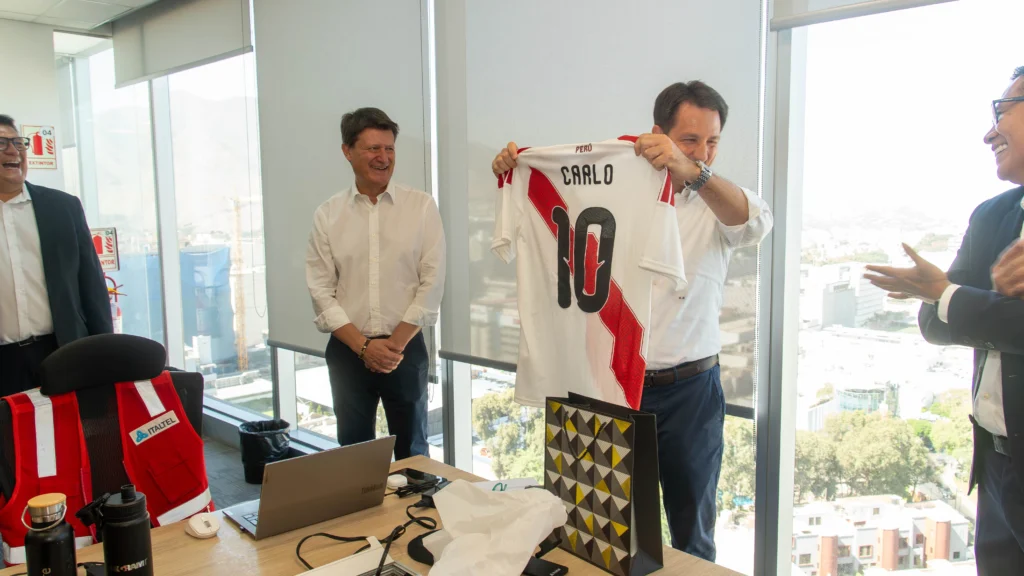 Business executive in a modern office presenting a personalized Peru national soccer team jersey with the name “CARLO” and number 10 during a corporate gathering or employee recognition event. Group of smiling business professionals in formal attire standing in a bright conference room with large city-view windows. Office desk with laptop, gift bag, reusable water bottles, and red safety vest visible in the foreground. Workplace celebration, corporate culture, team appreciation, international business meeting, and sports-themed employee gift presentation concept. Visible text on jersey includes “PERU,” “CARLO,” and “10.”