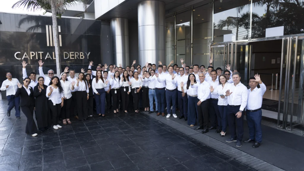 A large group of people in white shirts standing outside a building, waving and smiling.