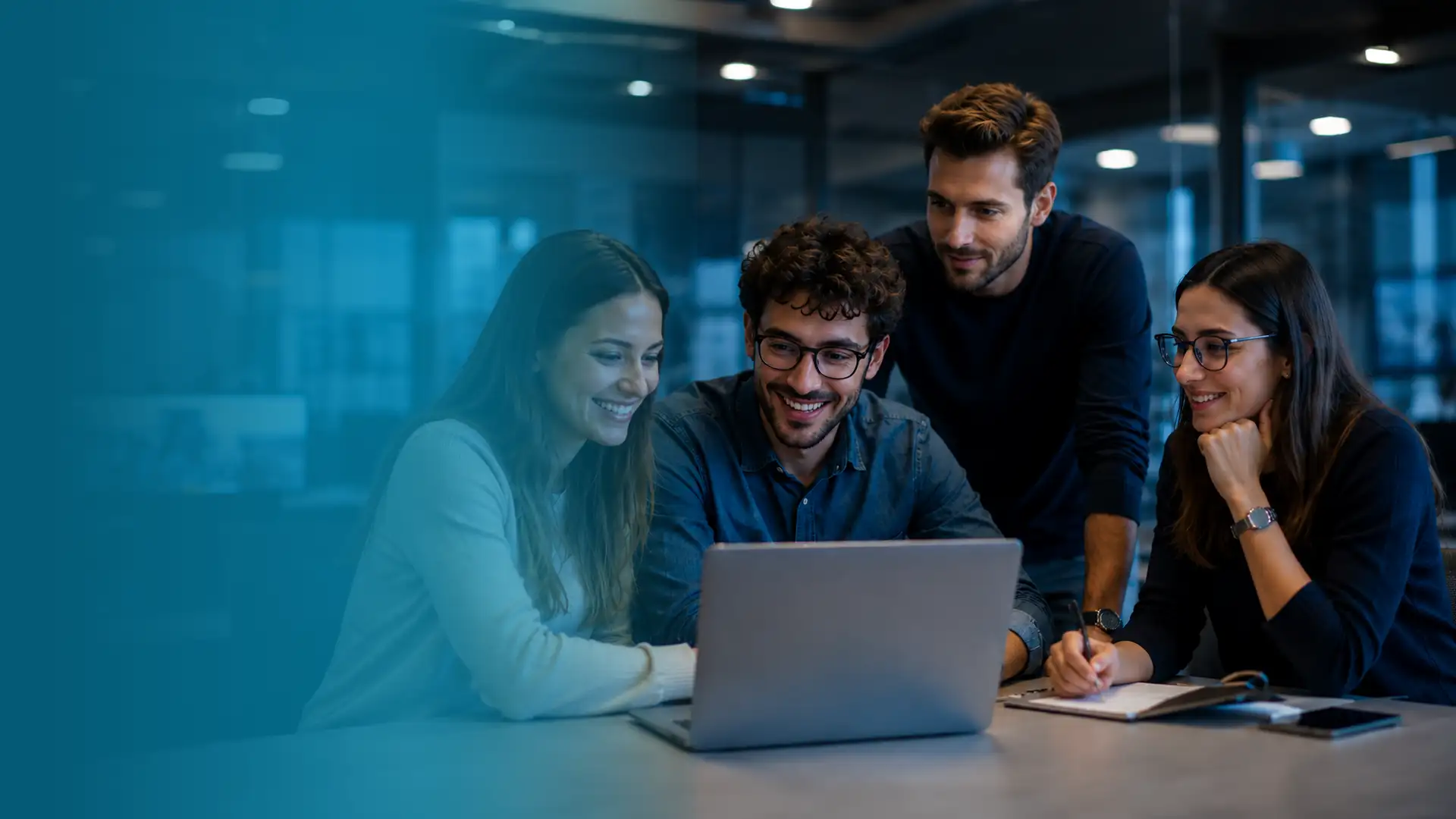A group of four smiling colleagues collaborate around a laptop in a dimly lit office .