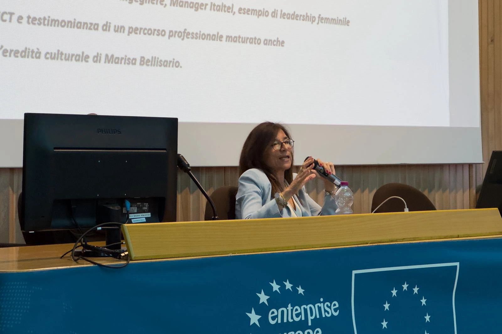 A woman - who is our colleague Loredana Tomasino - speaking at a conference, discussing female leadership in the ICT sector, with a computer and microphone in front of her.