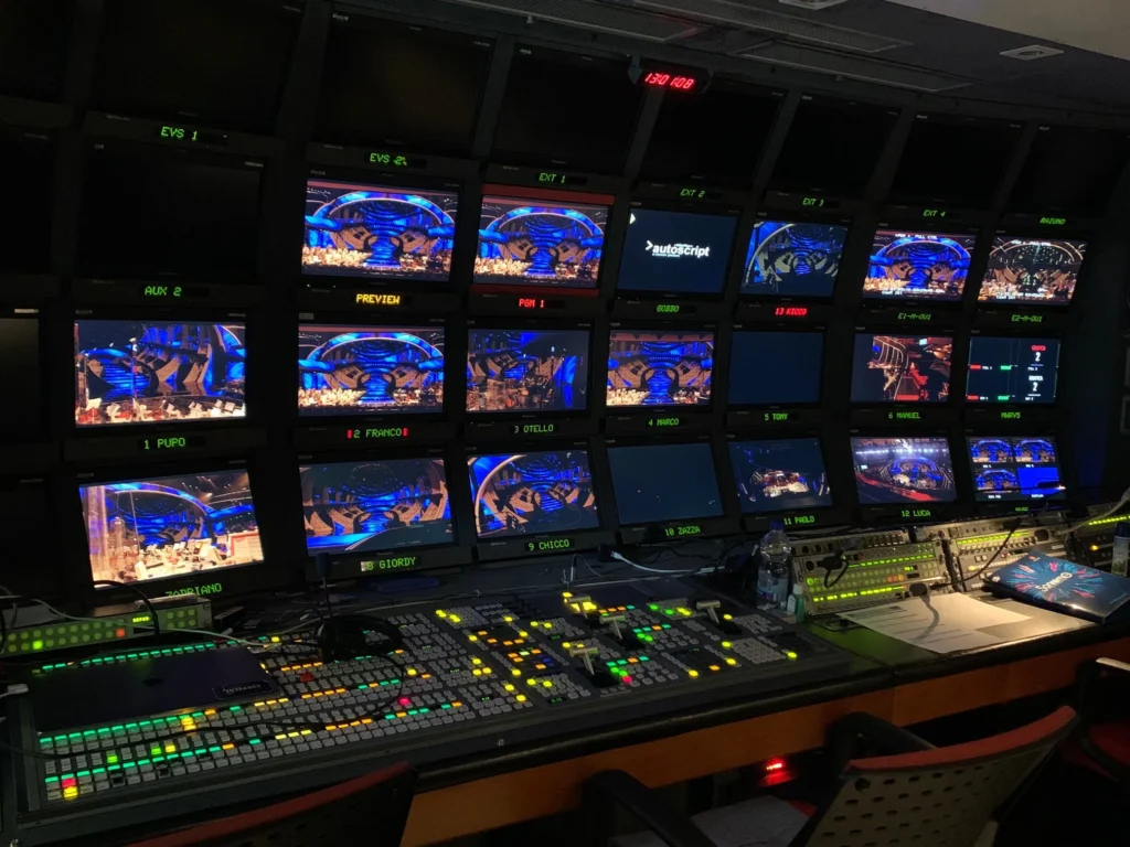 A television control room featuring multiple screens displaying a live event with colorful lighting and various control equipment.
