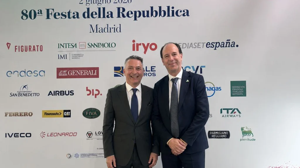 Alessandro Di Salvo & Ercole Rovida in suits smile in front of a backdrop featuring logos of various companies and the text "80ª Festa della Repubblica" in Madrid.